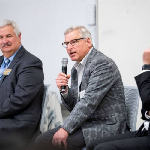 Three men hold microphones wearing official attires seem to address people in aroom.